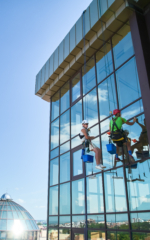 Meist 9 Industrial mountaineering. Male washers hanging on ropes and using cleaning tools while washing building windows. Two men window cleaners in safety helmets working together at high-rise building.
